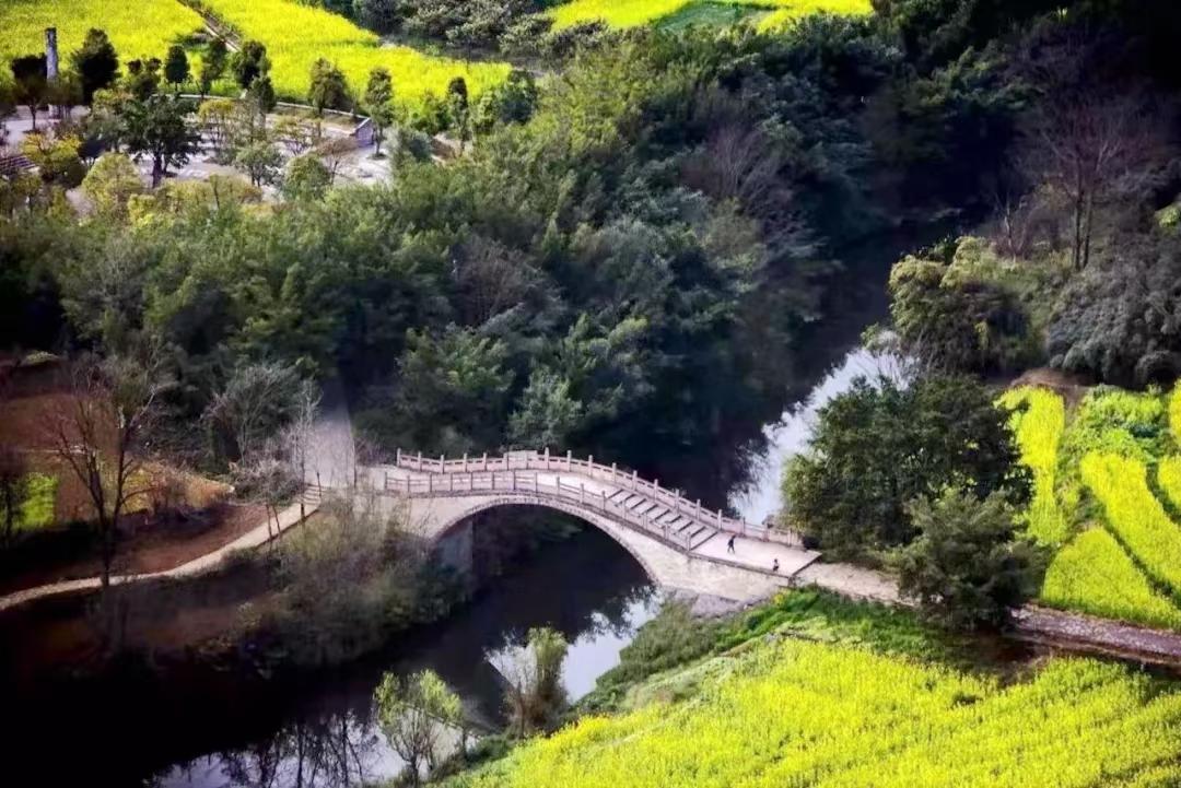 An old stone arch bridge over still water, with canola fields and karst peaks beyond in spring